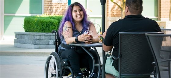 An adult in a wheelchair sits at a table and has a conversation with another adult