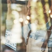 Closeup image of a storekeeper putting up a sign that says "Come In, We're Open" while smiling and looking out the window.