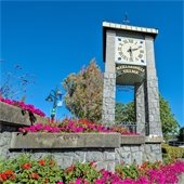 An image of Maillardville Village stone clocktower surrounded by pink and red flowers.