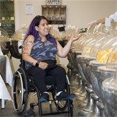 A young woman with purple hair and a camo-patterned shirt in a wheelchair looks at goods in circular bowls to purchase