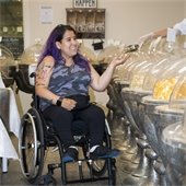 A young woman with purple hair and a camo-patterned shirt in a wheelchair looks at goods in circular bowls to purchase