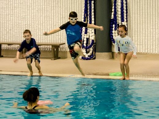 Four kids jumping into a an indoor pool towards an instructor