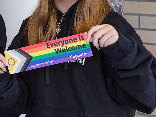 A youth holding up a sign that reads Everyone is Welcome with the Pride flag as the background. 