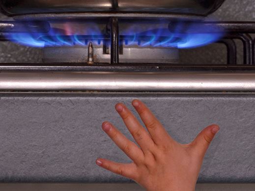 A small child's hand reaches up from the edge of the image towards a gas stove flame.