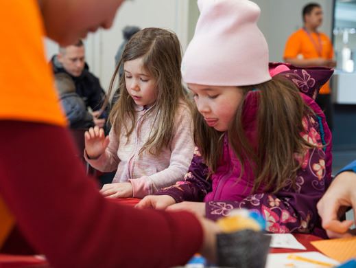 Two young children wearing winter clothes sit at a table and make crafts