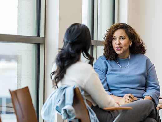 A woman in a blue sweater speaks to another woman in a cream shirt.