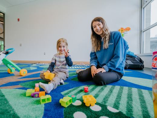 A City staff wearing a blue sweater sitting down next to a young girl in a recreation setting.
