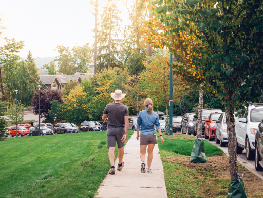 Photo of 2 people walking on a sidewalk with a cars parked along the side of the road